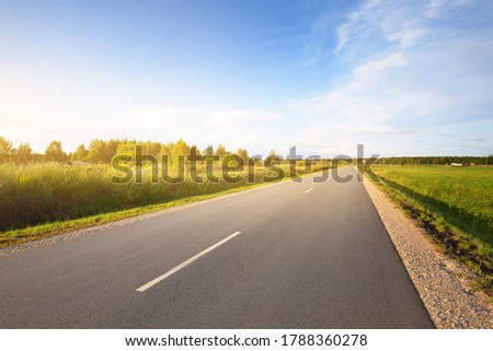 An empty highway (asphalt road) through the green country agricultural field, Latvia. Dramatic clouds after the rain. Idyllic summer rural scene. Dangerous driving, freedom concept