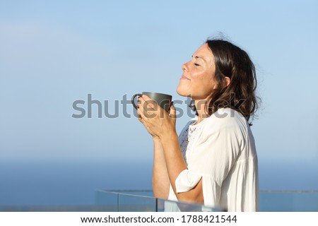 Side view portrait of a satisfied adult woman smelling coffee cup standing in a balcony on the beach Royalty-Free Stock Photo #1788421544
