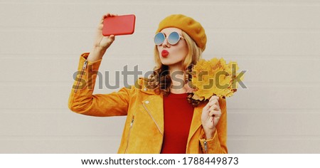 Autumn portrait of beautiful woman taking selfie picture by phone with yellow maple leaves wearing french beret over gray background
