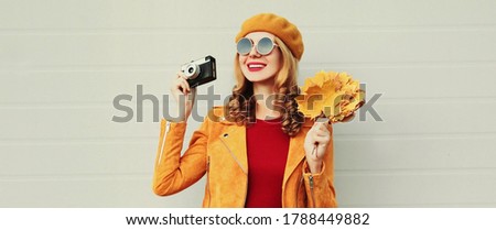 Autumn portrait of smiling young woman with retro camera and yellow maple leaves wearing french beret over gray background