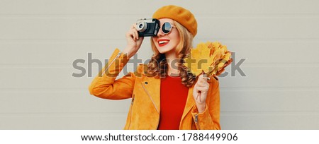 Autumn portrait of happy smiling woman with retro camera and yellow maple leaves wearing french beret over gray background