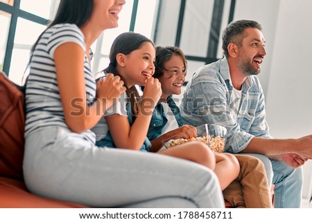 Beautiful mother and handsome father with their daughter and son spending time together at home. Sitting together on a couch, smiling eating popcorn and watching TV. Happy family concept.