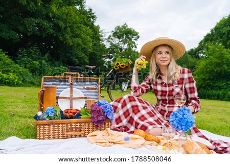 Girl in red checkered dress and hat sitting on white knit picnic blanket reading book and drinking wine. Summer picnic on sunny day with bread, fruit, bouquet hydrangea flowers. Selective focus.