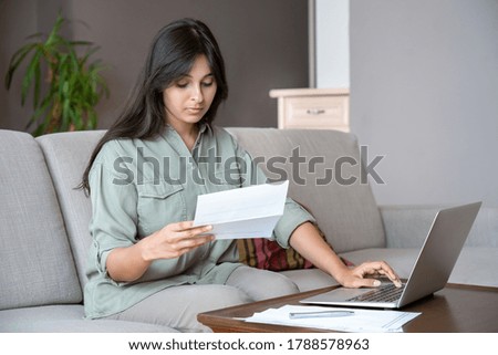 Young indian woman holding letter using laptop computer application paying bill online on website, managing account finances, calculating budget tax banking loan debt payment sitting on couch at home. Royalty-Free Stock Photo #1788578963