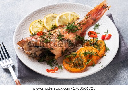 Baked sea bass with tomato pepper lemon and herbs in a ceramic plate on a gray concrete background. Perch fish cooked in the oven.
