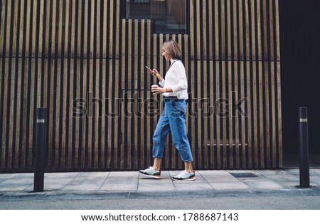 Side view of concentrated female pedestrian in stylish outfit text messaging on mobile phone while walking along pavement near contemporary building Royalty-Free Stock Photo #1788687143