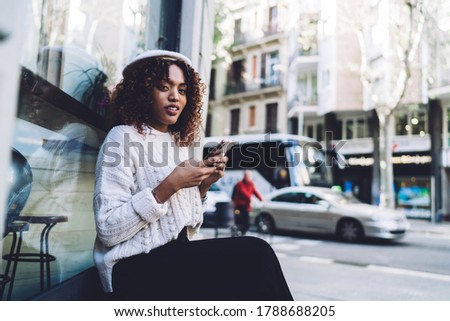 Side view of thoughtful African American female in casual clothes and hat sitting on wooden bench and browsing smartphone while resting on street and looking at camera