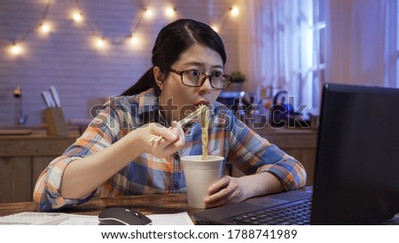 young japanese girl eats instant noodles and looks in laptop computer monitor. serious starving office lady worker working at home in night kitchen and having bedtime snack with unhealthy fast food