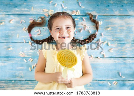 Funny little girl with sprinkled lips and freckles on color background with colorful candies. Kid holding a caramel yellow candy on a stick. Happy childhood concept