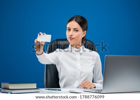 Beautiful, young businesswoman sitting at her desk before a laptop, showing a blank slip of paper, isolated on blue background.