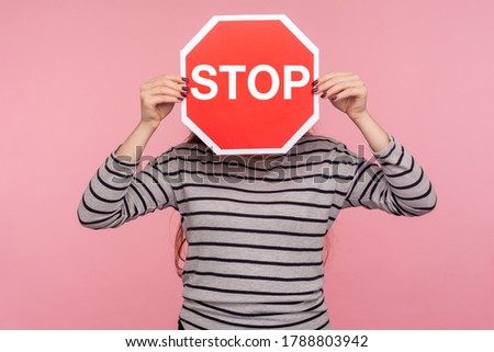 Way prohibited! Portrait of anonymous person in striped sweatshirt covering face with Stop symbol, holding red traffic sign and warning of ban, forbidden access. indoor studio shot, pink background