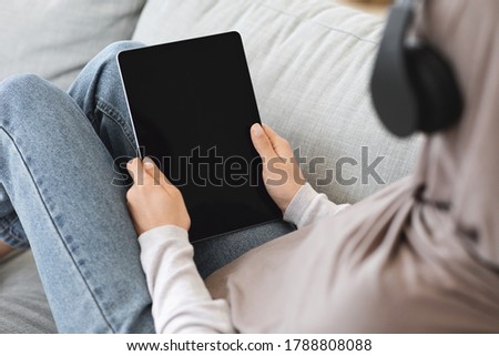 Mockup Image Of Digital Tablet With Black Screen In Hands Of Muslim Woman Relaxing On Couch At Home, Over Shoulder View