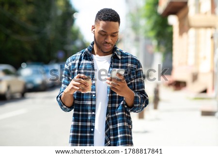 Good-looking young black hipster messaging online on his cell phone, walking down the street with coffee cup Royalty-Free Stock Photo #1788817841