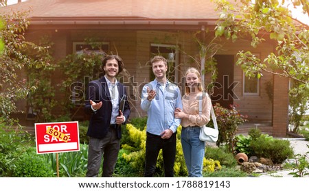 Happy realtor and millennial couple standing on front of beautiful country house, panorama