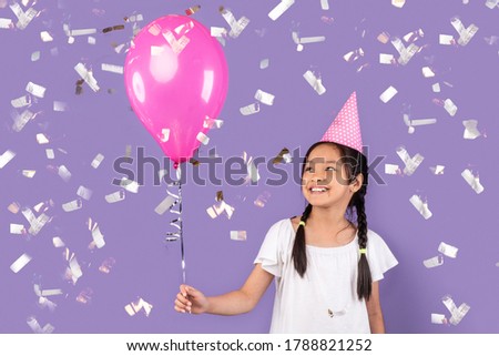 Kid's Birthday Party. Happy Japanese Girl Holding Pink Balloon And Wearing Hat Celebrating B-Day Under Falling Confetti Over Purple Background.