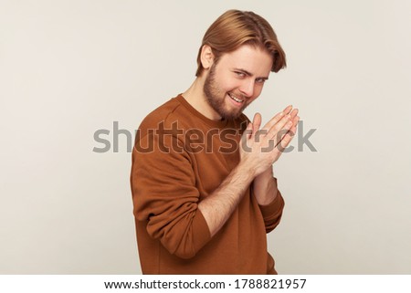 Portrait of sneaky sly scheming man with beard wearing sweatshirt rubbing palm as having cunning evil idea, devious plan in mind, thinking revenge. indoor studio shot isolated on gray background Royalty-Free Stock Photo #1788821957