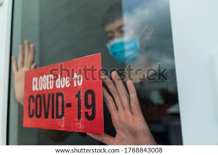 Young asian man setting up red sign hanging at the glass door of a shop saying (Closed due to covid-19). Coronavirus concept. Respiratory protection. Quarantine at home.