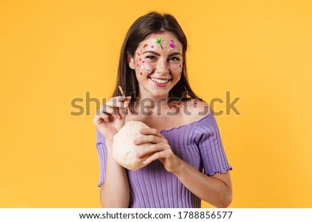 Image of young laughing woman with stickers on face drinking fresh coconut water isolated over yellow background