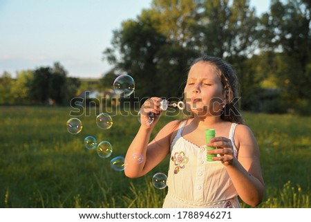 Portrait of little cute girl which blows soap bubbles smiling. Outdoors on a background of green grass and forest.