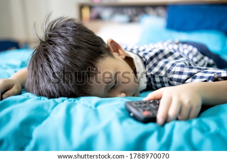Asian kid boy is sleeping while watching cartoons online on TV with remote control in hand,little child fell asleep studying online,abnormal drowsiness,Narcolepsy disease in children,sleep disorders 