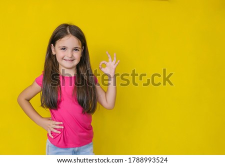 A little cute happy girl looking at the camera and showing a sign of approval, OK, on a yellow background.The child smiles. Space for your text.
