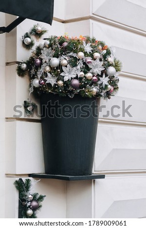 Decorative pot in the form of a black bucket with a Christmas bouquet of flowers, fir branches, toys and shiny tinsel on gray wall. The facade of the house near the entrance to the store.