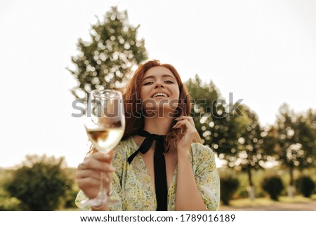 Joyful red haired lady with black bandage on her neck in modern green dress smiling, holding glass with drink and looking into camera outdoor..