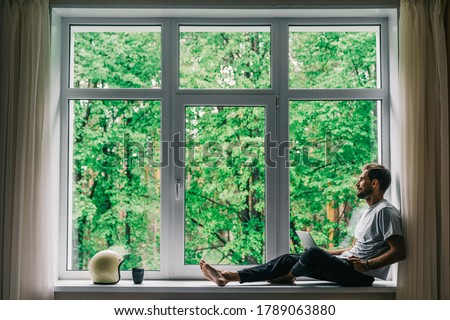 Corona virus - staying and work at home (self-isolation, self-quarantine). A self-isolated handsome man sits alone in front of a panoramic window with a laptop. Online shopping, home work, freelance. Royalty-Free Stock Photo #1789063880