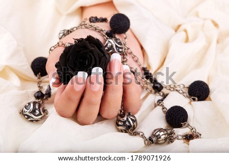 Hand with long artificial French manicured nails holding black rose flower and necklace