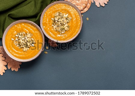 Pumpkin soup with pumpkin seeds in bowls and dry autumn leaves on a dark gray background top view. Free space for text. Copy space. Flat lay. Autumn food. American cuisine.