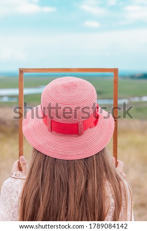 Girl in pink straw hat holds empty wooden picture frame in hands on nature landscape background faceless. Portrait of a woman with long hair posing on photo back view. Creative idea concept.