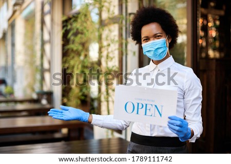 Happy black waitress with face mask and glover hodling open sign while reopening care during coronavirus epidemic. 
