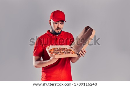 Young bearded delivery man in red uniform looks in surprise at a box of pizza isolated on gray background.