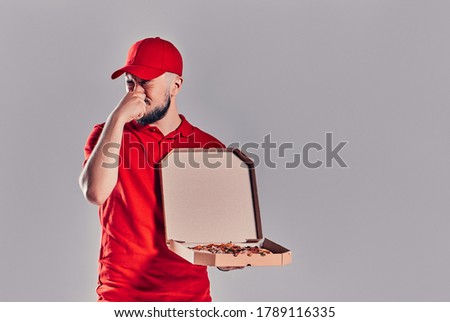 Young bearded delivery man in a red uniform with pizza covers his nose with his hand from an unpleasant smell isolated on gray background.