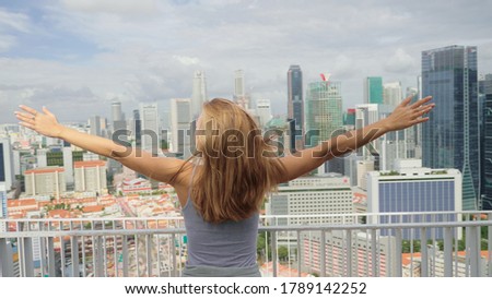 A happy tourist girl holds up her hands and enjoys the stunning view of the skyscrapers in Singapore on a warm summer day. Travel destination, adventure, success and exploration concept.