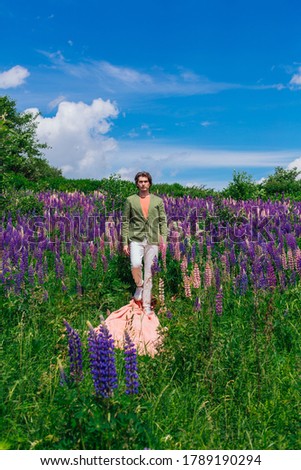Tall handsome man in a green jacket walking on a pink cloth in lupine flowers field and enjoing the beauty of nature. Man surrounded by purple and pink lupines.