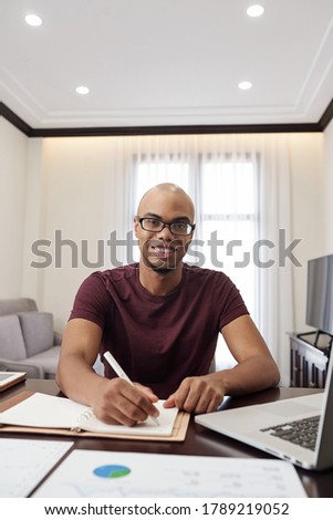 Positive bald young African-American man sitting at desk at home and filling planner