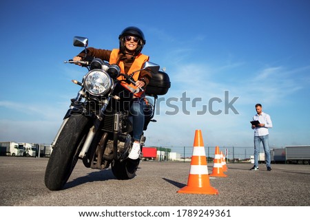 Motorcycle school of driving. Female driver with helmet taking motorcycle lessons and practicing ride. In background traffic cones and instructor with checklist rating and evaluating the ride.  Royalty-Free Stock Photo #1789249361