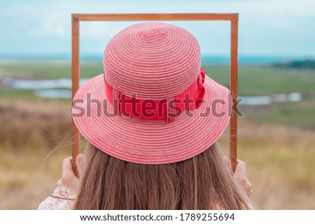 Girl in pink straw hat holds empty wooden picture frame in hands on nature landscape background. Portrait of a woman with long hair posing back view. Creative idea concept.