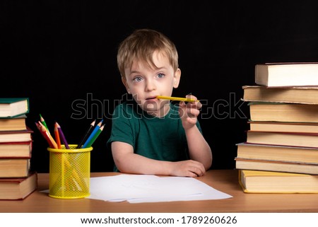 a three-year-old boy sits at a table with books and gnaws a colored pencil. home leisure. the child draws pictures. black wall on background. isolate.