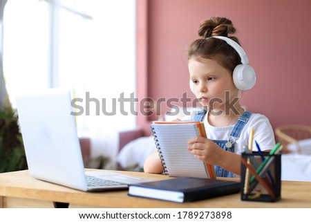 Little girl in headphones sit at desk writing in notebook studying online do exercises at home, little child handwrite prepare homework on quarantine, have web class or lesson indoors.
