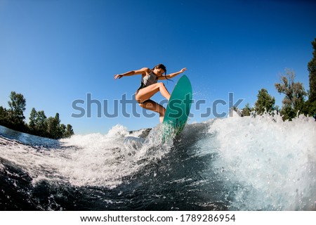 young wet woman in gray swimsuit energetically balancing on wave on wakesurf board. Clear blue sky and trees in the background.
