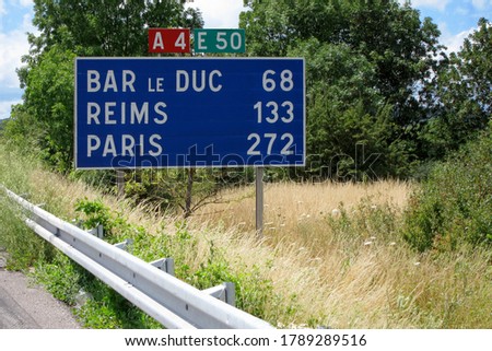 A road sign to the towns of Bar le Duc, Reims and Paris, 272 km away, on an abandoned highway among green trees and grass on the wayside. Sunny day. Copy space.                               