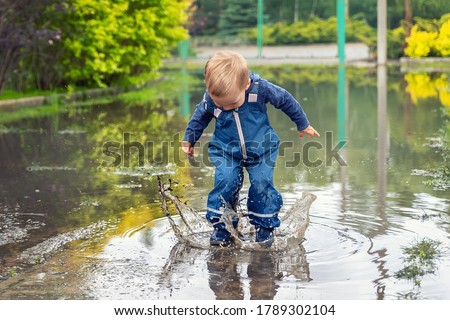 Little cute playful caucasian blond toddler boy enjoy have fun playing jumping in dirty puddle wearing blue waterproof pants and rubber rainboots at home yard street outdoor. Happy childhood concept Royalty-Free Stock Photo #1789302104