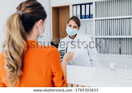 Young female practitioner or nurse with face protective mask working at clinic reception desk. She is helping to patient to make contactless payment for medical services.