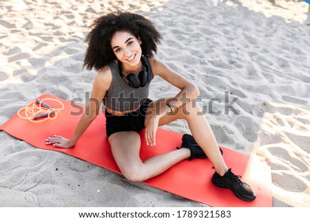 A beautiful woman with an afro hair sits on the gymnastic mat on the beach and looks at camera with a smile. An athletic beautiful biracial girl in sportswear with headphones is doing outdoor workout