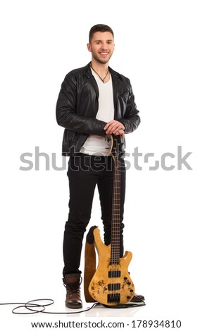 Smiling guitarist in black leather jacket stands with bass guitar. Full length studio shot isolated on white.