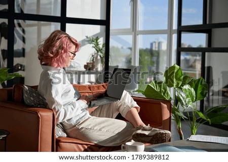 Stylish creative hipster young woman, teen girl with pink hair using pc laptop computer browsing internet work learn communicate online sit on sofa in modern home office lit with morning sunshine.