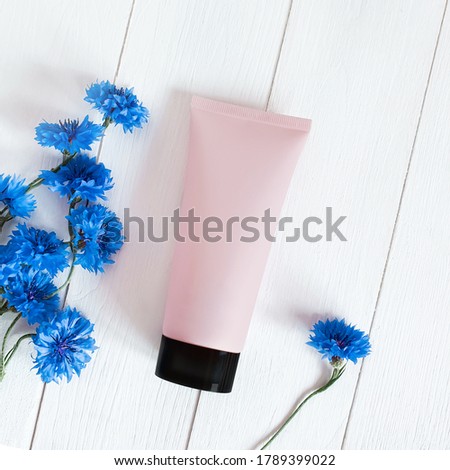 Top view of mock-up pink squeeze bottle plastic tube with black cap and blue cornflowers on a white wooden background. Natural organic spa cosmetics concept. Flatlay, square photography.