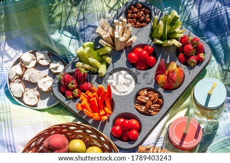 Summertime healthy picnic with fresh vegetables, nuts and fruits served in the muffin pan, refreshing drink in mason jar and some snak on blue cloth in the garden.